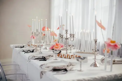 Elegant gedeckter Esstisch für ein Event, perfekt für ein stilvolles Modelshooting in Solingen. Der Tisch ist mit hohen weißen Kerzen in silbernen Haltern, rosa Blumen und makellosem weißen Geschirr geschmückt. Ordentlich gefaltete Servietten und poliertes Silberbesteck runden das edle Ambiente ab.