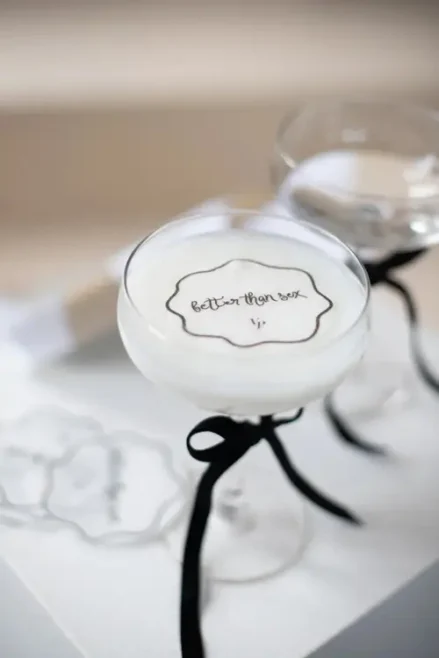 In einem schicken Modelshooting-Setting in Solingen ziert ein Glas mit schaumiger weißer Flüssigkeit ein Coupe-Glas, das mit einer schwarzen Schleife am Stiel verziert ist. Die Oberfläche ziert ein exquisites Design mit dem Text „Better than Sex
