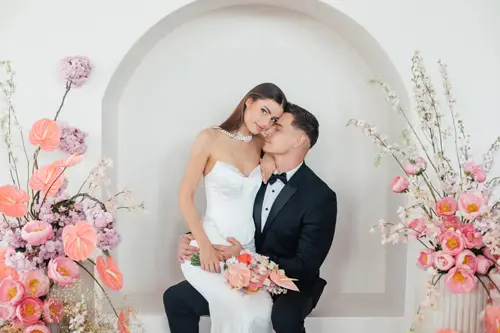 Eine Braut im weißen Kleid und ein Bräutigam im schwarzen Smoking sitzen in einer dekorativen Nische zusammen. Umgeben von rosa Blumen, die eine zauberhafte Kulisse für ihren intimen Moment schaffen, küsst der Bräutigam die Braut sanft auf die Stirn. Die perfekte Kulisse für ein Modelshooting in Solingen.