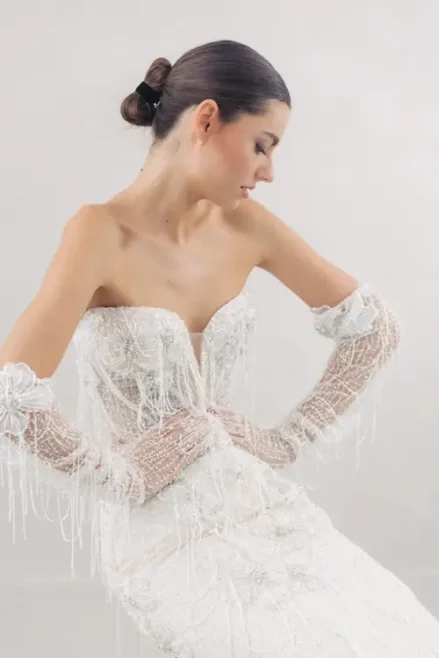 Bei einem Modelshooting in Solingen trägt eine Frau ein trägerloses weißes Kleid mit aufwendiger Perlenstickerei und dazu passende, ellenbogenlange Handschuhe. Ihr Haar ist zu einer glatten Hochsteckfrisur gestylt, während sie vor einem hellen Hintergrund seitlich nach unten blickt.