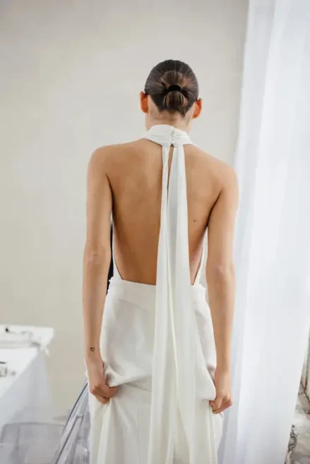 In minimalistischer Umgebung mit hellen Wänden und drapierten Vorhängen blickt ein Model mit Dutt abgewandt in ein rückenfreies Neckholder-Kleid mit fließendem, weißem Schal. Dieser fesselnde Moment fängt die Essenz eines Modelshootings in Solingen ein.