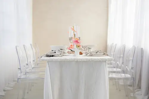 Ein minimalistisches Esszimmer-Setup, ideal für ein Modelshooting in Solingen, besteht aus einem langen, mit einer weißen Tischdecke bedeckten Tisch. Transparente Stühle säumen beide Seiten, während weiße Teller, Besteck und rosa Blumen ein elegantes Herzstück bilden. Weiche weiße Vorhänge hängen friedlich im Hintergrund.