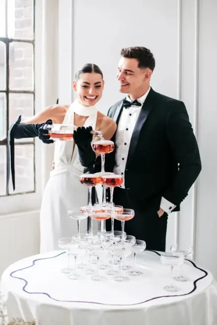 Bei einem Modelshooting in Solingen schenkt eine Frau in weißem Kleid und schwarzen Handschuhen elegant Champagner in einen Turm aus Gläsern ein. Neben ihr steht ein Mann im Smoking mit einem warmen Lächeln. Das Tageslicht, das durch das Fenster des hellen Raumes strömt, lässt die beiden in ihren Bann ziehen.