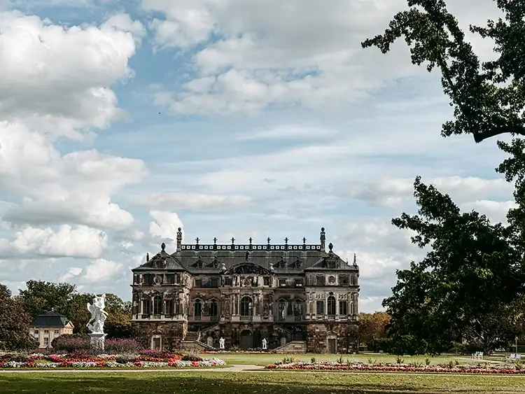 Ein historisches, verziertes Herrenhaus aus Stein steht in einem gepflegten Garten mit bunten Blumenbeeten und Statuen, umgeben von Bäumen unter einem teilweise bewölkten Himmel.