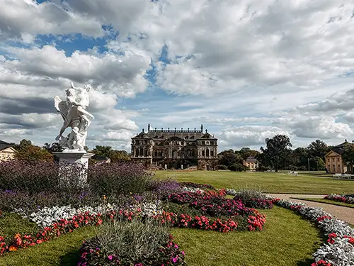 Garten mit bunten Blumenbeeten und einer weißen Statue im Vordergrund, der zu einem großen historischen Gebäude unter einem teilweise bewölkten Himmel führt.