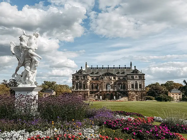 Im Hintergrund steht ein prächtiges historisches Gebäude mit verschnörkelter Architektur, während eine weiße Marmorstatue und farbenfrohe Blumenbeete den Vordergrund unter einem teilweise bewölkten Himmel füllen.