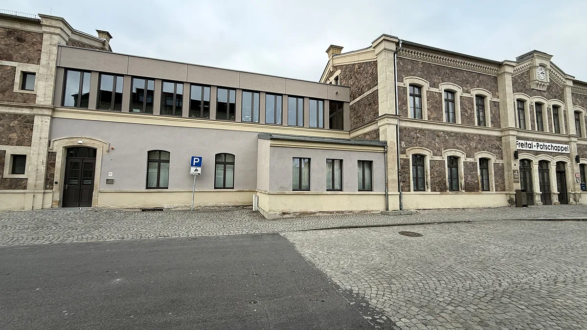 Ein historisches Postgebäude mit einer Steinfassade steht neben einem modernen Anbau mit großen Fenstern; davor sind eine Kopfsteinpflasterstraße und ein Parkschild zu sehen.
