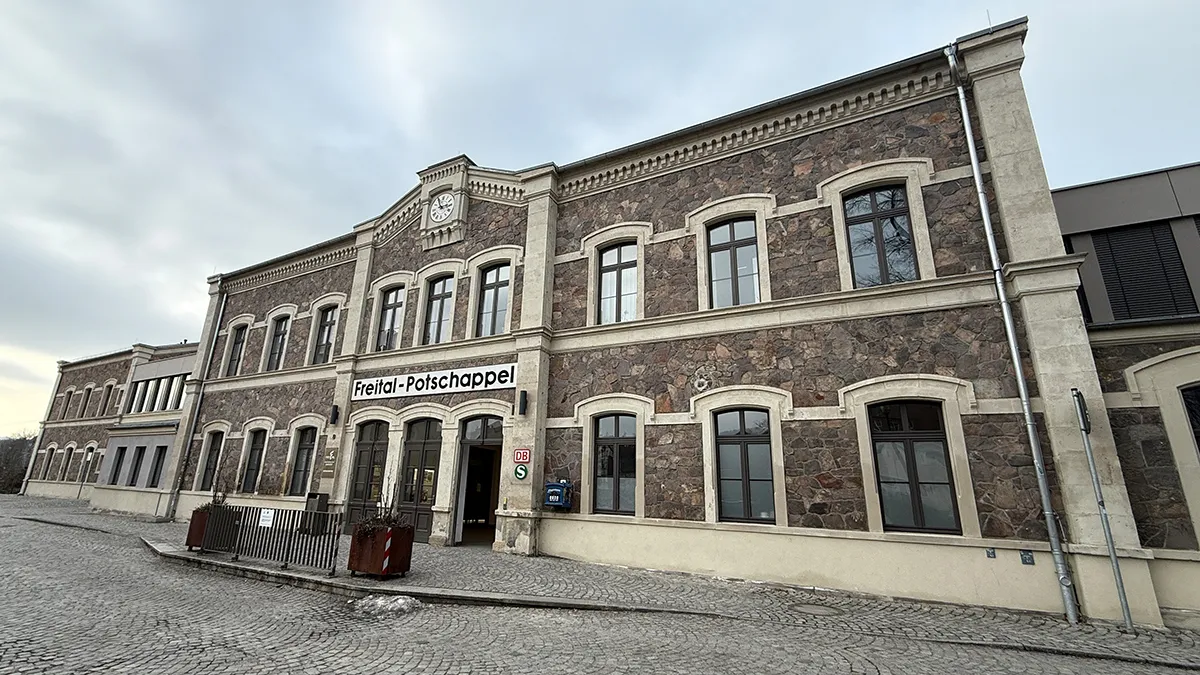 Ein historisches Bahnhofsgebäude aus Stein mit Rundbogenfenstern und einer Uhr über dem Eingang, beschriftet mit "Freital-Potschappel". Der Himmel ist bedeckt und die Straße davor ist mit Kopfsteinpflaster gepflastert.