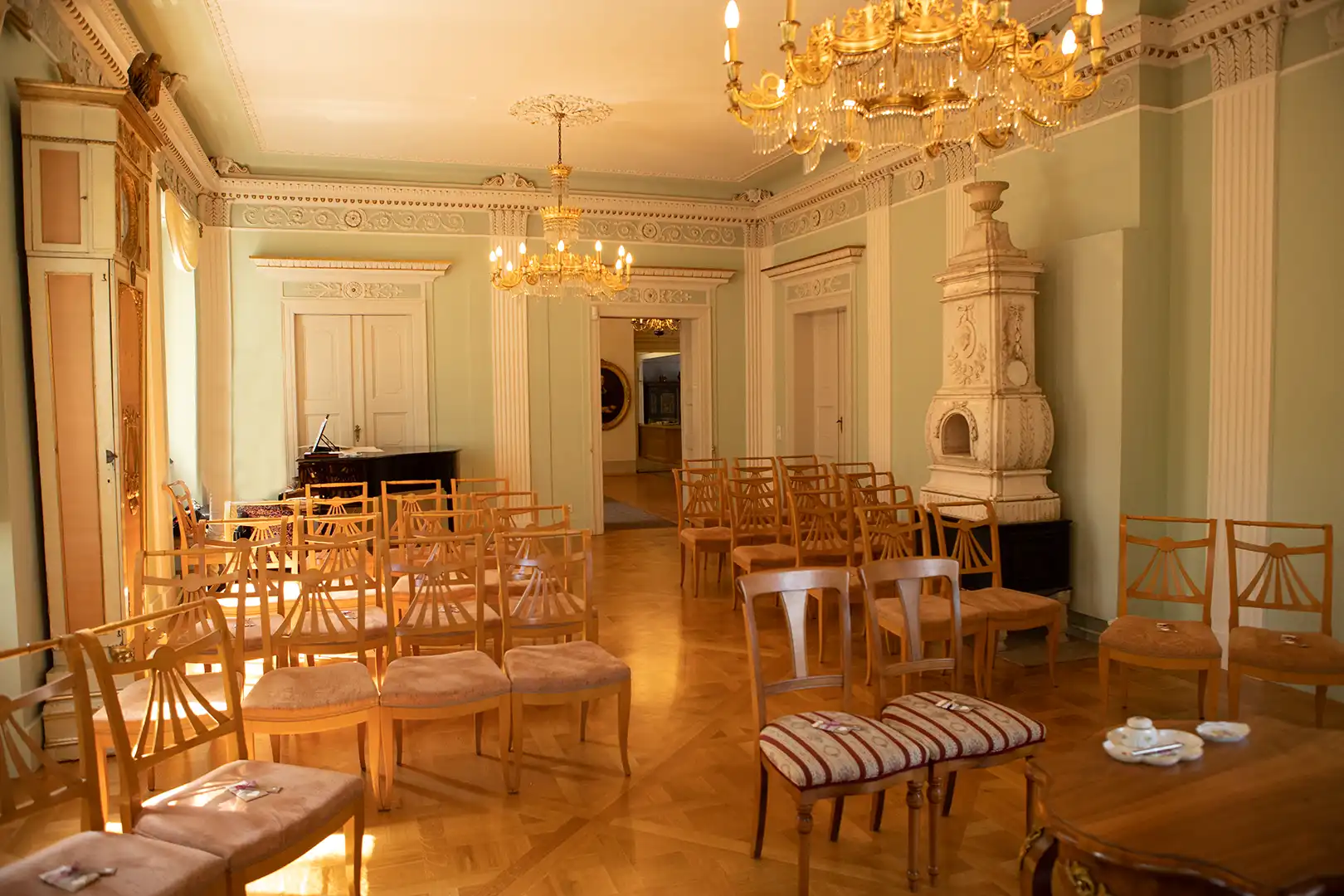 Ein heller, eleganter Raum mit kunstvollen Kronleuchtern an der Decke. Reihen von Holzstühlen stehen vor einem Flügel in der Ecke. Dieser bezaubernde Raum auf Schloss Burgk eignet sich perfekt, um besondere Momente festzuhalten, wie Melanie Kunert Fotografie in ihrer Arbeit als Hochzeitsfotografin Freital eindrucksvoll beweist.