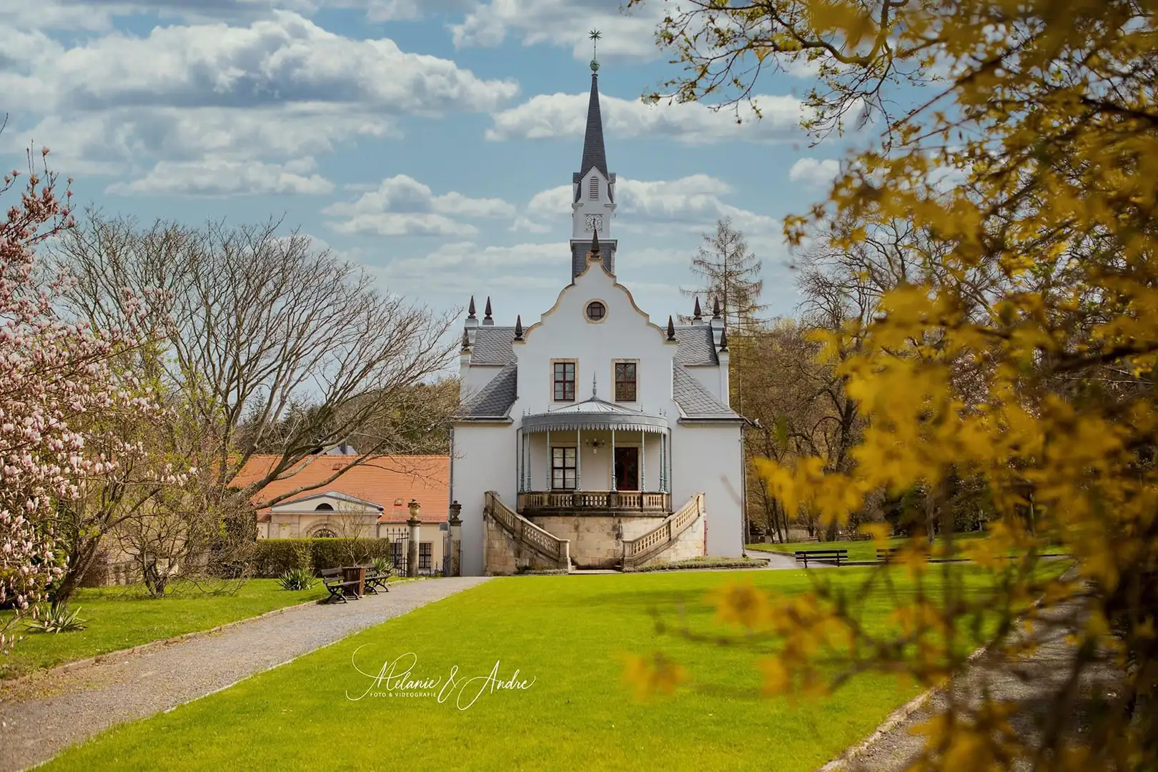 Ein malerisches kleines Schloss mit einem hohen Turm inmitten eines üppigen grünen Rasens und leuchtend blühender Bäume erinnert an Schloss Burgk. Hochzeitsfotograf Freital fängt die ruhige Frühlingsstimmung unter einem klaren blauen Himmel mit flauschigen Wolken ein.