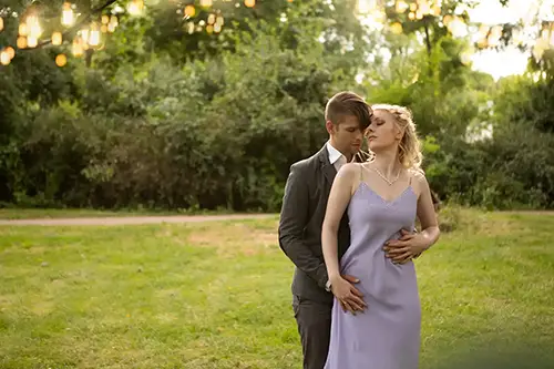 Ein Paar steht in einem Park, umgeben von Grün. Der Mann umarmt die Frau von hinten, während beide nach unten blicken. Die Frau trägt ein lavendelfarbenes Kleid und der Mann einen grauen Anzug. Sonnenlicht fällt durch die Bäume und schafft eine sanfte, romantische Atmosphäre.