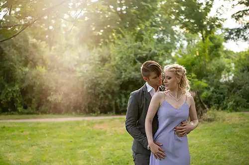 Ein Paar steht in einem Park, umgeben von Grün. Der Mann umarmt die Frau von hinten, während beide nach unten blicken. Die Frau trägt ein lavendelfarbenes Kleid und der Mann einen grauen Anzug. Sonnenlicht fällt durch die Bäume und schafft eine sanfte, romantische Atmosphäre.