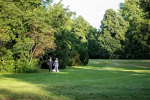 Ein Paar geht Hand in Hand einen von üppigen grünen Bäumen gesäumten Weg in einem Park entlang. Sonnenlicht fällt durch das Laub und wirft Schatten auf das Gras. Die Szene ist ruhig und malerisch.