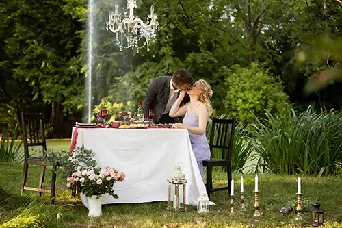 Ein Paar küsst sich an einem romantisch gedeckten Tisch im Freien unter einem Kronleuchter. Der Tisch ist mit einem weißen Tuch, Blumen, Kerzen und verschiedenen Speisen geschmückt. Sie sind von üppigem Grün in einer Gartenkulisse umgeben.