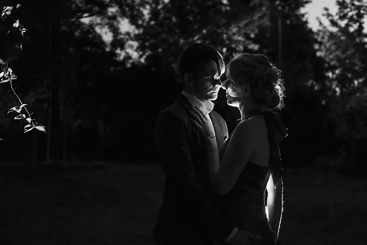 Silhouette eines Paares, das sich im Dunkeln eng gegenübersteht, mit sanftem Gegenlicht, das ihre Profile hervorhebt. Die Szene ist intim und romantisch, mit verschwommenen Bäumen im Hintergrund.