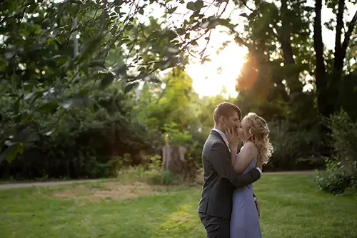Ein Paar umarmt sich bei Sonnenuntergang in einem üppigen Garten. Der Mann im Anzug und die Frau im langen Kleid teilen einen intimen Moment, während das Sonnenlicht durch die Bäume fällt und die Szene in einen warmen Schein taucht.
