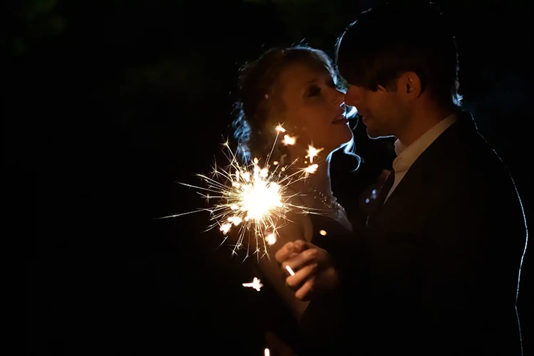 Ein Paar küsst sich im Dunkeln, beleuchtet von einer Wunderkerze in der Hand des Mannes. Ein sanftes Licht sorgt für einen schwachen Schimmer hinter ihnen und hebt ihre Silhouetten in der Dunkelheit hervor – wunderschön eingefangen von der Expertin für Verlobungsshooting Dresden, Melanie Kunert Fotografie.
