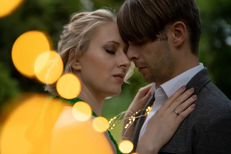Ein Paar umarmt sich in einer Gartenkulisse, umgeben von sanftem Bokeh-Licht. Die Frau trägt ein grünes Kleid und hat hochgesteckte Haare, während der Mann einen Anzug trägt und mit Lichterketten umhüllt ist. Sie scheinen einen intimen Moment zu teilen.