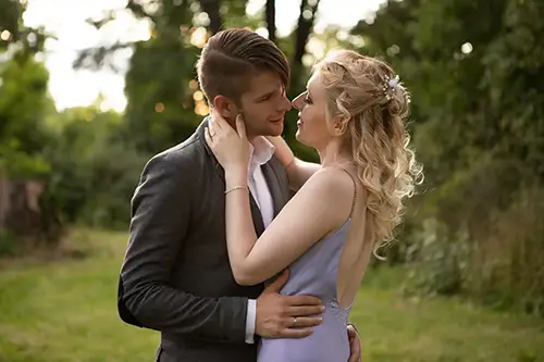 Ein Paar umarmt sich im Freien, steht auf einer Wiese mit Bäumen im Hintergrund. Die Frau, die ein hellviolettes Kleid trägt, hat ihre Arme um den Hals des Mannes gelegt. Der Mann trägt eine dunkle Jacke und hält sie an der Taille. Beide schauen sich in die Augen.