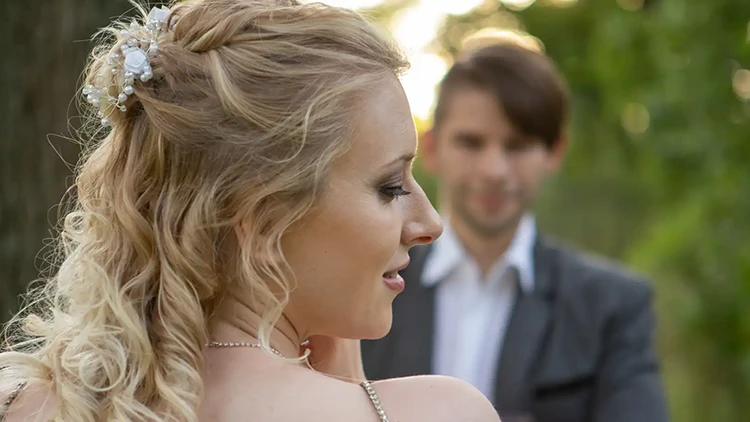 Eine Frau mit lockigem blondem Haar, das mit kleinen weißen Blumen geschmückt ist, blickt im Freien über ihre Schulter. Im unscharfen Hintergrund steht ein Mann im Anzug und lächelt sanft. Üppiges Grün umgibt sie und suggeriert eine natürliche Umgebung.
