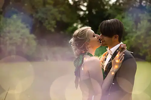 Ein Paar umarmt sich in einer Gartenkulisse, umgeben von sanftem Bokeh-Licht. Die Frau trägt ein grünes Kleid und hat hochgesteckte Haare, während der Mann einen Anzug trägt und mit Lichterketten umhüllt ist. Sie scheinen einen intimen Moment zu teilen.