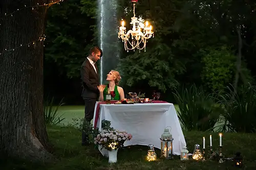 Ein Paar genießt ein romantisches Abendessen im Freien. Ein Kronleuchter hängt über einem mit einem weißen Tuch bedeckten Tisch, der mit Kerzen und Blumen geschmückt ist. Der Mann steht neben dem Tisch, während die Frau sitzt. Im Hintergrund ist ein Wasserfall zu sehen.