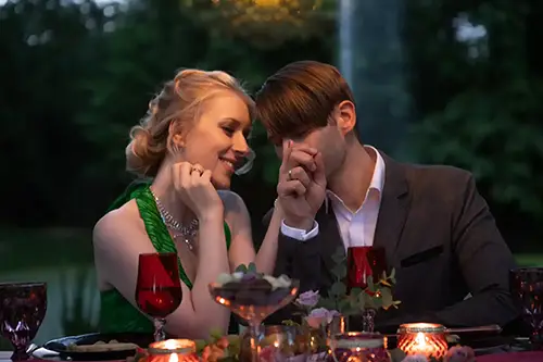 Ein Paar sitzt in romantischer Atmosphäre an einem Esstisch im Freien und hält Rotweingläser in den Händen. Die Frau trägt ein grünes Kleid und der Mann einen grauen Anzug. Der Tisch ist mit Kerzen, Blumen und Häppchen dekoriert. Der Hintergrund ist verschwommenes Grün.