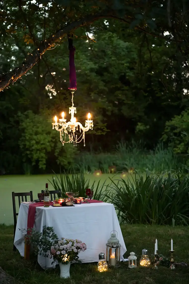 Ein romantisches Outdoor-Dinner erwartet Sie mit einem weiß gedeckten Tisch und Holzstühlen, perfekt für ein *Verlobungsshooting in Dresden*. Die Szene, geschmückt mit einem hängenden Kronleuchter, Blumenarrangements und dekorativen Kerzen, wird von üppigen grünen Bäumen und Gras eingerahmt.
