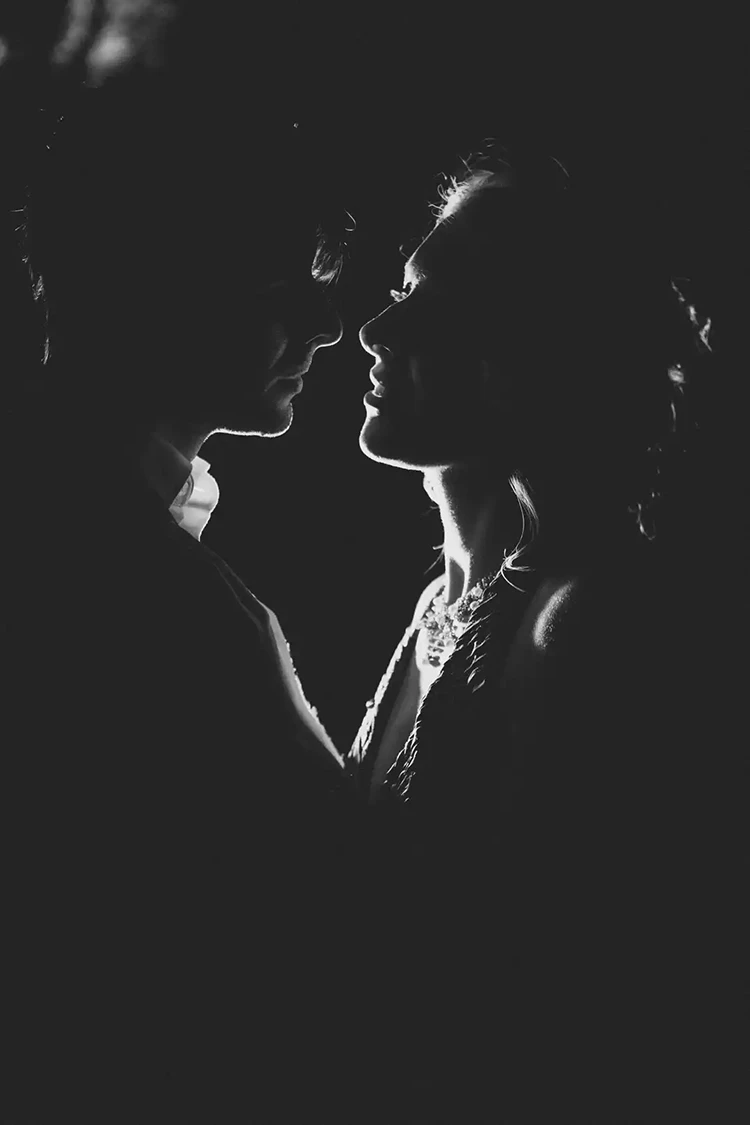 Eine schwarzweiße Silhouette eines Paares, das sich eng gegenübersteht, wobei weiches Licht das Gesicht der Frau hervorhebt. Ihre Ausdrücke deuten auf Intimität hin, vor einem dunklen Hintergrund.