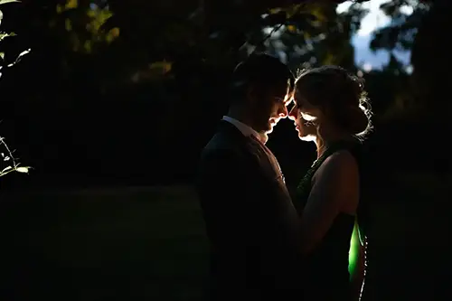 Silhouette eines Paares, das sich im Dunkeln eng gegenübersteht, mit sanftem Gegenlicht, das ihre Profile hervorhebt. Die Szene ist intim und romantisch, mit verschwommenen Bäumen im Hintergrund.