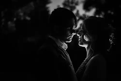 Silhouette eines Paares, das sich im Dunkeln eng gegenübersteht, mit sanftem Gegenlicht, das ihre Profile hervorhebt. Die Szene ist intim und romantisch, mit verschwommenen Bäumen im Hintergrund.