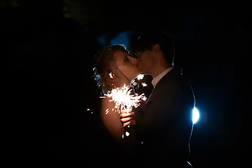 Ein Paar küsst sich im Dunkeln, beleuchtet von einer Wunderkerze in der Hand des Mannes. Ein sanftes Licht sorgt für einen schwachen Schimmer hinter ihnen und hebt ihre Silhouetten in der Dunkelheit hervor – wunderschön eingefangen von der Expertin für Verlobungsshooting Dresden, Melanie Kunert Fotografie.