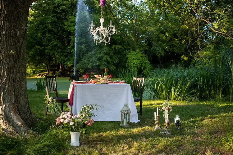 Ein romantisches Outdoor-Dinner erwartet Sie mit einem weiß gedeckten Tisch und Holzstühlen, perfekt für ein *Verlobungsshooting in Dresden*. Die Szene, geschmückt mit einem hängenden Kronleuchter, Blumenarrangements und dekorativen Kerzen, wird von üppigen grünen Bäumen und Gras eingerahmt.