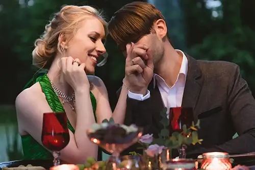 Ein Paar sitzt draußen an einem Tisch bei Kerzenlicht. Die Frau im grünen Kleid lächelt herzlich, während der Mann im grauen Anzug ihr zärtlich die Hand küsst. Rotweingläser und Blumenschmuck sorgen für eine intime Atmosphäre, die Hochzeitsfotografin Dresden-Melanie Kunert Fotografie bei ihrem Verlobungsshooting Dresden perfekt eingefangen hat.