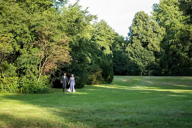 Ein Paar geht Hand in Hand einen von üppigen grünen Bäumen gesäumten Weg in einem Park entlang. Sonnenlicht fällt durch das Laub und wirft Schatten auf das Gras. Die Szene ist ruhig und malerisch.