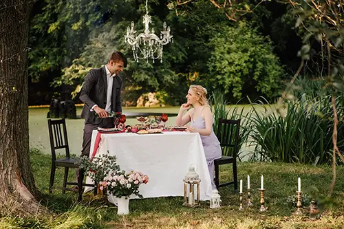 Ein Paar genießt ein elegantes Picknick im Freien an einem Teich. Der Mann im Anzug schenkt der Frau, die in einem lavendelfarbenen Kleid sitzt, Wein ein. Der Tisch ist mit Blumen und Kerzen geschmückt, und darüber hängt ein kunstvoller Kronleuchter, umgeben von üppigem Grün.