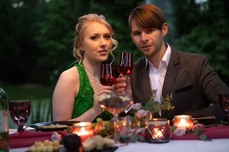 Ein Paar sitzt in romantischer Atmosphäre an einem Esstisch im Freien und hält Rotweingläser in den Händen. Die Frau trägt ein grünes Kleid und der Mann einen grauen Anzug. Der Tisch ist mit Kerzen, Blumen und Häppchen dekoriert. Der Hintergrund ist verschwommenes Grün.