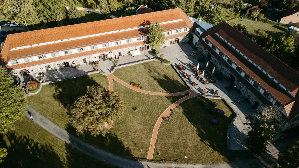 Luftaufnahme eines großen Anwesens, das sich perfekt für eine Hochzeit am Gut am See eignet, mit zwei langen Gebäuden mit roten Dächern, die einen gepflegten Rasen, sich kreuzende Gehwege und üppige Grünflächen umgeben.